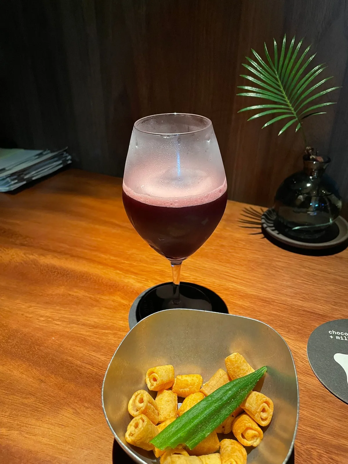 A deep red cocktail in a wine glass with the bar snack of crisps and okra at Chocolate+Milk