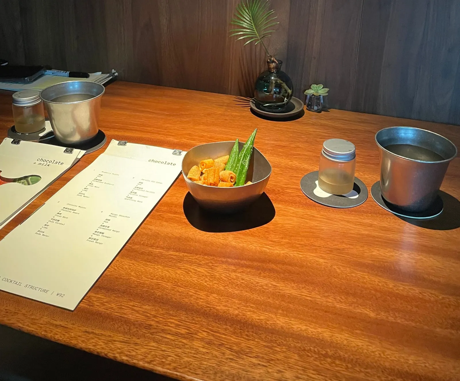 The Chocolate+Milk cocktail menu on a wooden table at the bar in Beijing, with crisps, okra and two tin cups of water