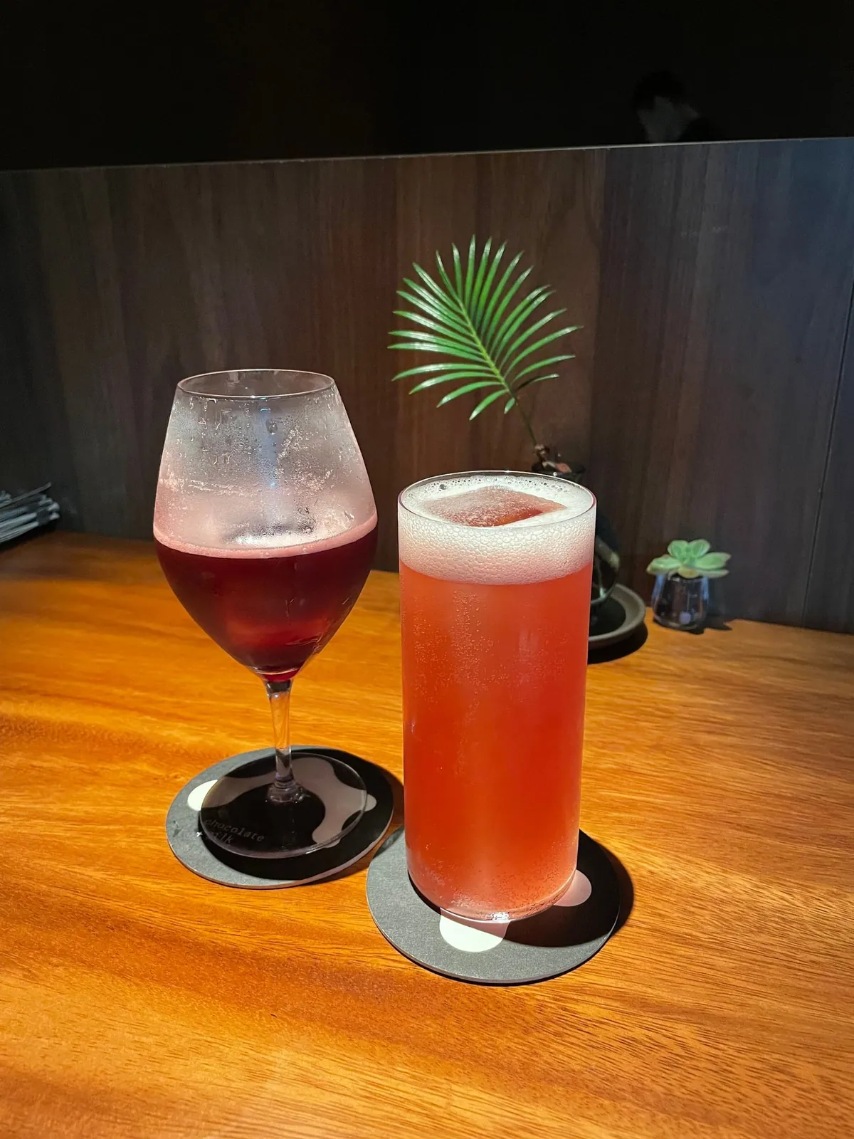 A dark red cocktail in a wine glass next to a pink, foam-topped highball at Chocolate+Milk